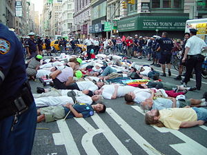 Picture of a street protest scene, with people lying down in a street, surrounded by police, onlookers, and stores in a big city.