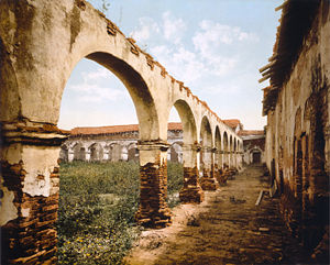 (PD) Photo: William Henry Jackson / Library of Congress A photocrom print of the ruins of an arcade that forms Mission San Juan Capistrano's cuadrángulo, circa 1899. Plants grow from the top of the colonnade, and rubble is piled against a wall.