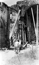 (PD) Photo: A. Yorba Several men work on the side of an arched structure, with one man atop a scaffolding, another below him, and yet another in the foreground carrying two buckets. The remains of two arched structures can be seen on either side of the construction area, while a patch of overgrown grass sits in the foreground, at right.